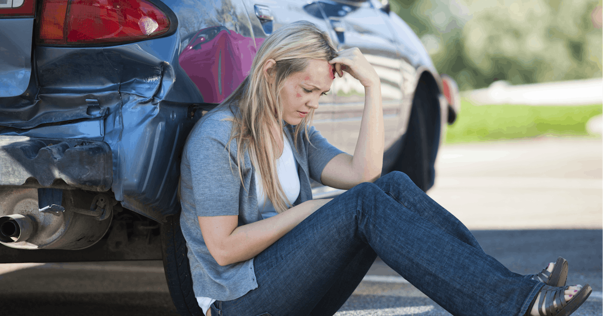 injured person sits on ground beside damaged car